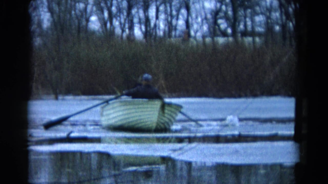 Man rowing a pale wooden boat across icy water with reeds and bare trees behind him.