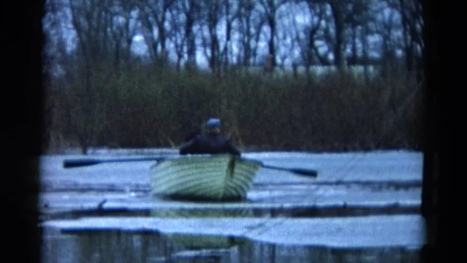 A distant adult rower in a pale boat crosses icy water in Wauconda, Illinois, with bare shoreline trees in the background.