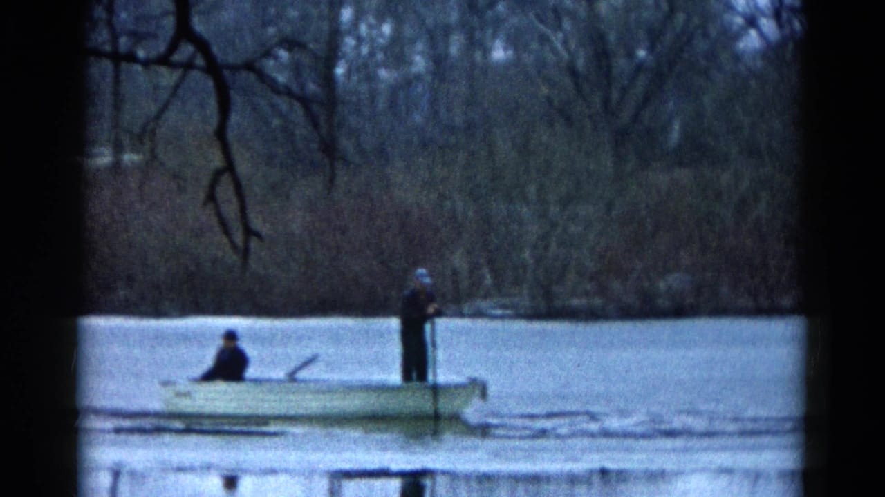 Small rowboat with two people crossing water in front of a wooded shoreline.
