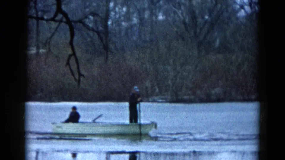 A distant 1961 home-movie view shows two people in a small rowboat on a Wauconda lake, framed by bare branches and reeds.