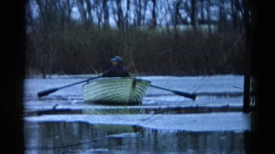 A person rows a pale boat on a Wauconda lake in 1961 while another passenger sits low, with reeds and cool-toned water behind them.