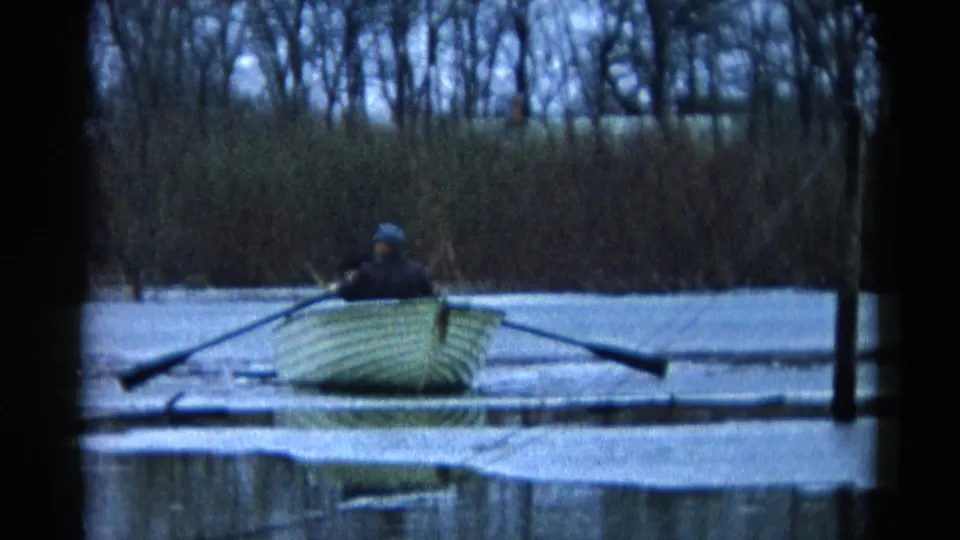 The rowboat is seen from behind on the Wauconda lake, with one person rowing, another seated, and bare trees across the shoreline.