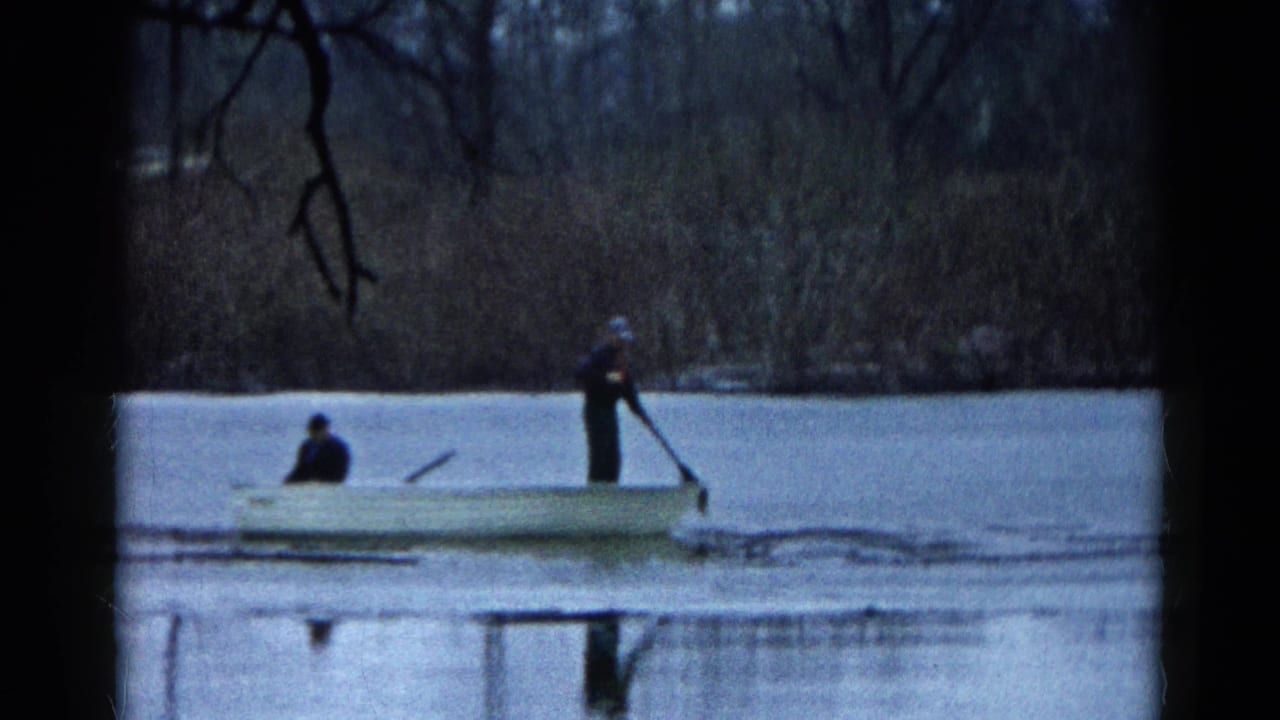 Two people in a small boat on an icy lake, with bare trees in the background.