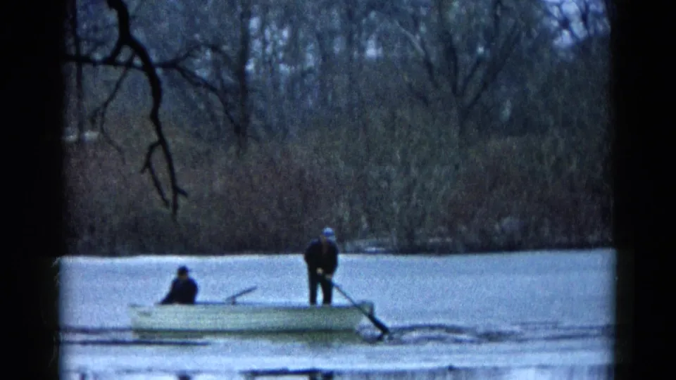 1961 8mm frame of two people in a small boat on an icy Wauconda lake, seen through dark tree branches.