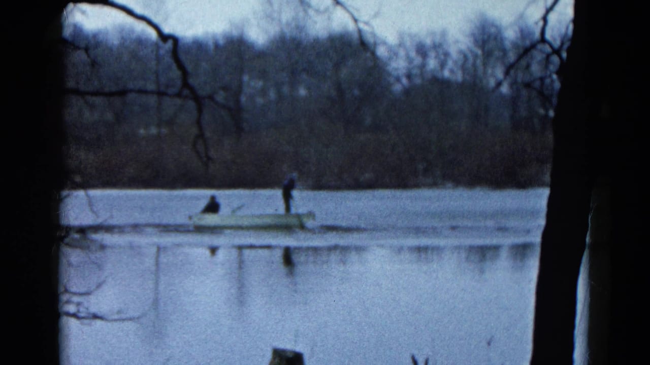 Two people in a small rowboat on a calm lake, seen through bare tree branches.
