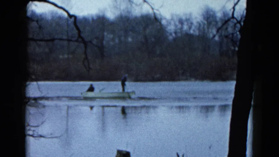 1961 8mm home-movie frame of two distant people in a small rowboat on a lake in Wauconda, Illinois, framed by dark branches.