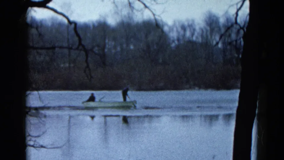 Two unidentified people travel in a small rowboat on a calm Wauconda lake, with bare trees and dark film edges visible.