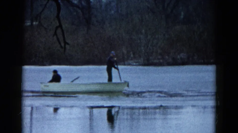 Closer 8mm frame of two unidentified people in a small rowboat on a Wauconda lake, one seated and one standing with an oar.