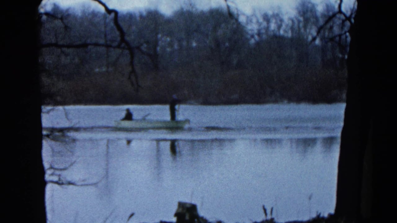 1961 Wauconda, Illinois home-movie frame showing two distant people in a small boat on a blue-gray lake with bare trees beyond.