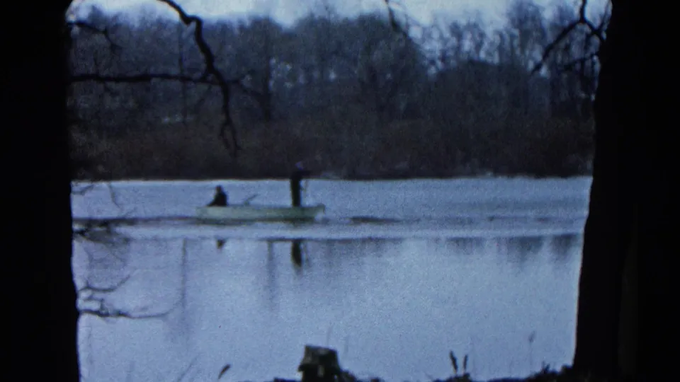 1961 Wauconda, Illinois home-movie frame showing two distant people in a small boat on a blue-gray lake with bare trees beyond.
