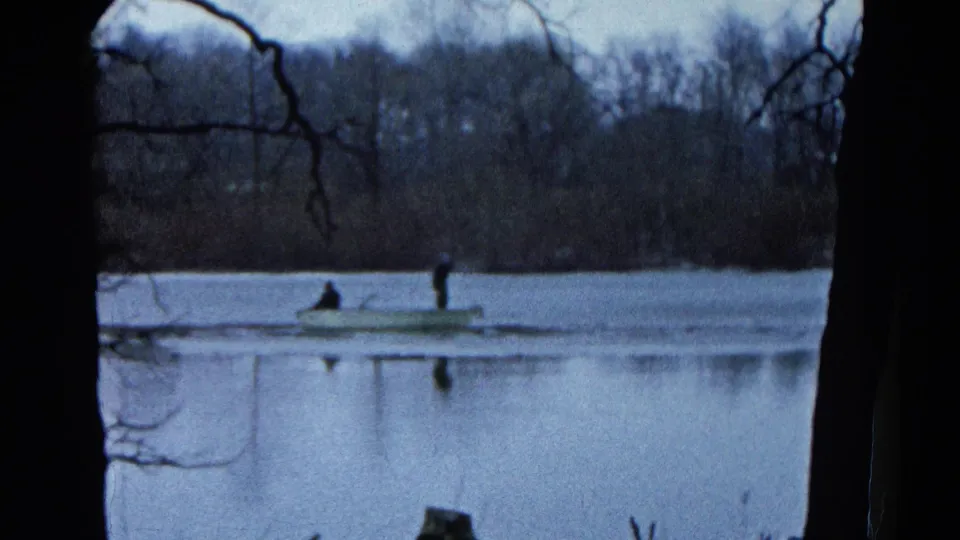 1961 Wauconda, Illinois frame of two people in a small lake boat, with foreground trees and a wooded shoreline in soft focus.