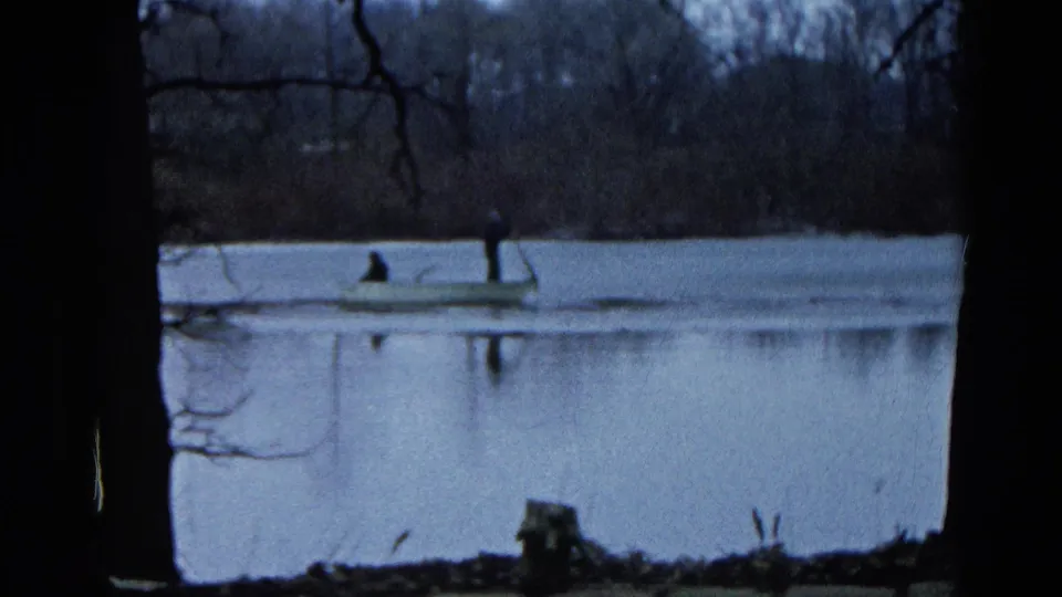 Distant 1961 lake scene in Wauconda, Illinois, with two people in a rowboat seen between dark foreground trees.
