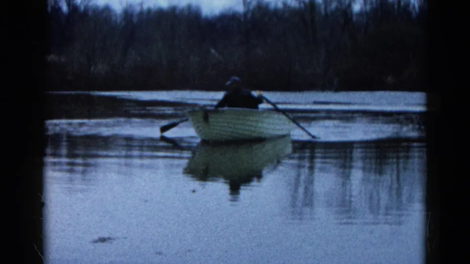 Home-movie frame from Wauconda, Illinois in 1961 showing a dark-clothed rower in a pale boat on reflective water at dusk.