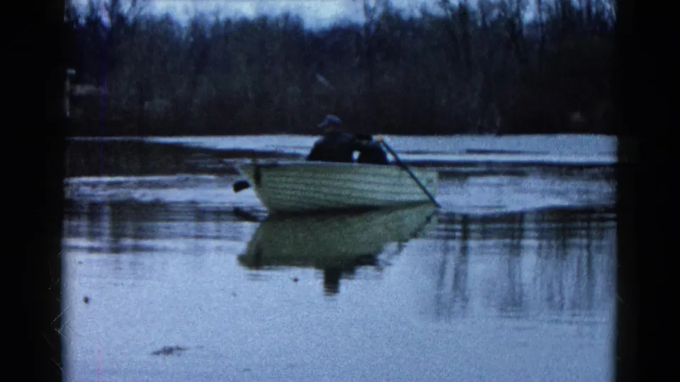 1961 Wauconda, Illinois frame with the rower and small boat shifted across the lake, trees and water reflections behind.
