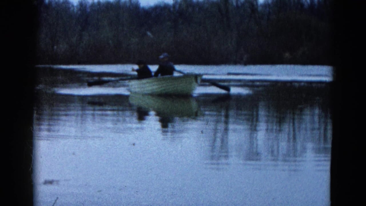 1961 Wauconda, Illinois 8mm frame showing two people in a light rowboat moving across calm water near a dark tree-lined shore.