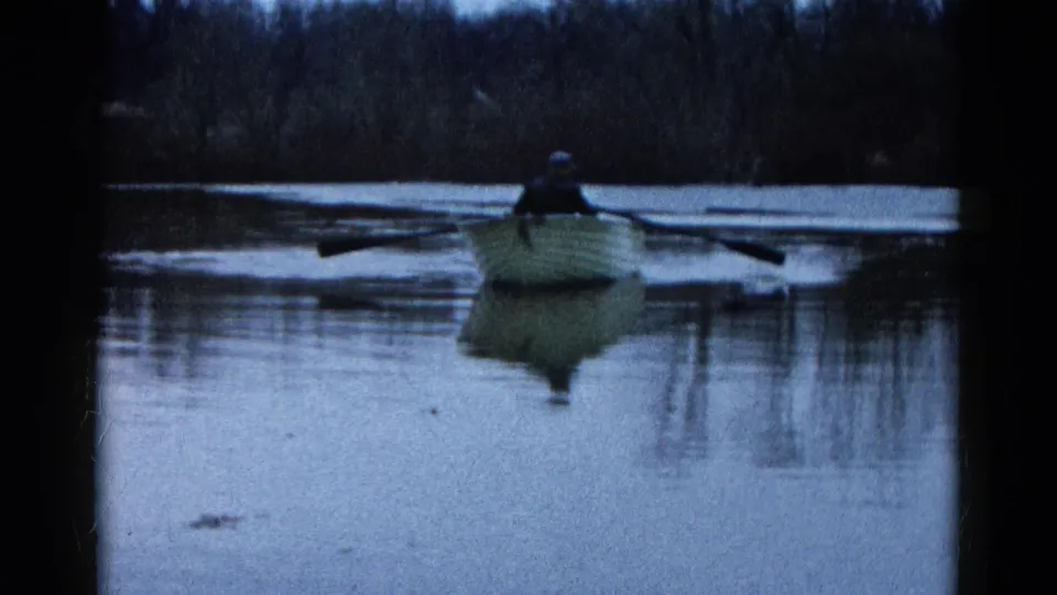 1961 Wauconda, Illinois frame with a centered rowboat, two people, broad oars, and rippled reflections below a wooded shore.