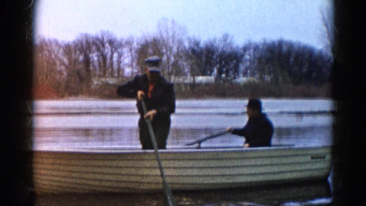 In Wauconda, Illinois, 1961 home-movie footage, two men in dark coats and hats push a yellow rowboat from the near shore.