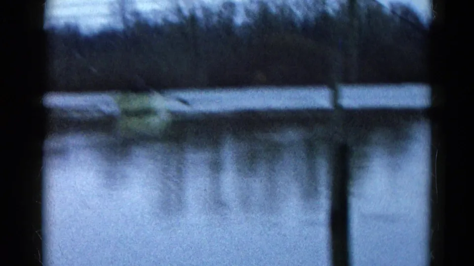 A soft, blue-shifted 8mm view shows the yellow rowboat across the Wauconda pond with bare trees and a vertical foreground edge.