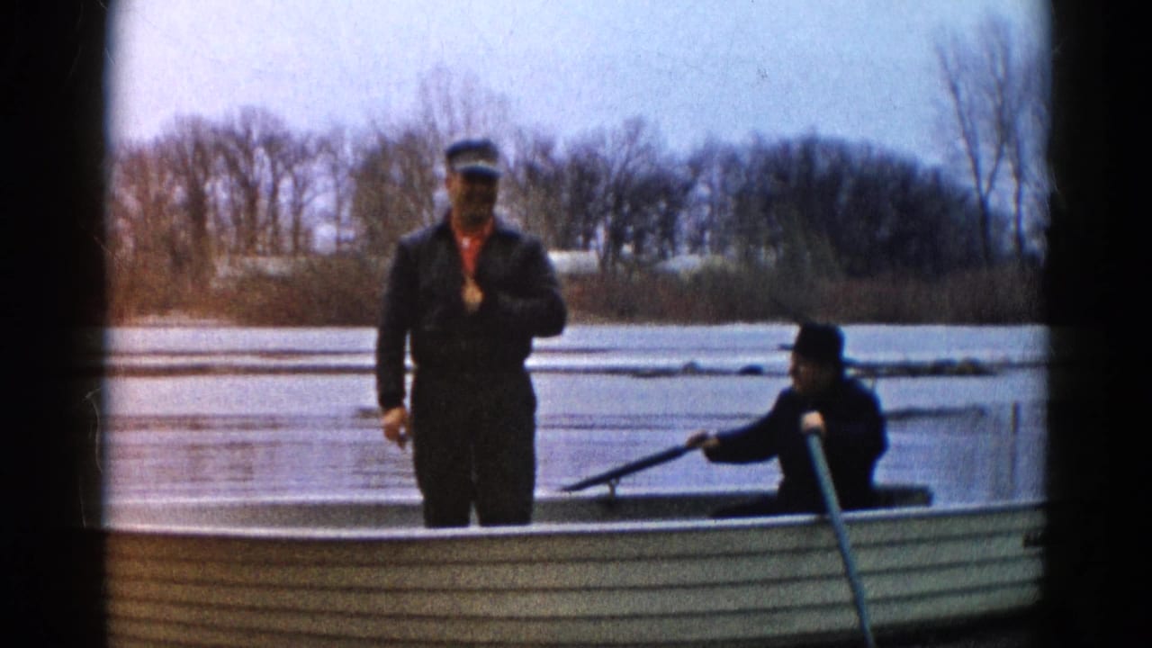 Two men in a rowboat on calm water, one standing near the bow and one seated with an oar.