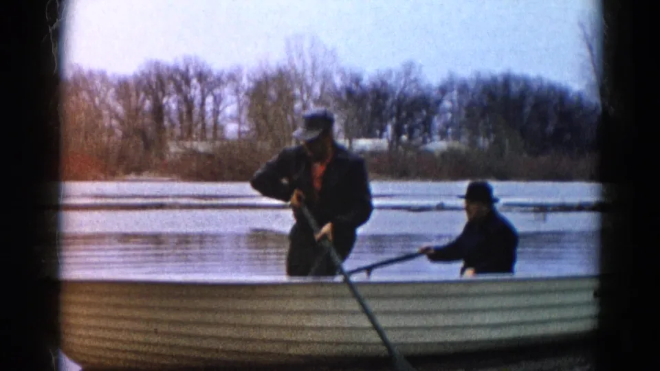 1961 Wauconda, Illinois frame with one man standing in a rowboat and another seated rowing near a bare lakeshore.