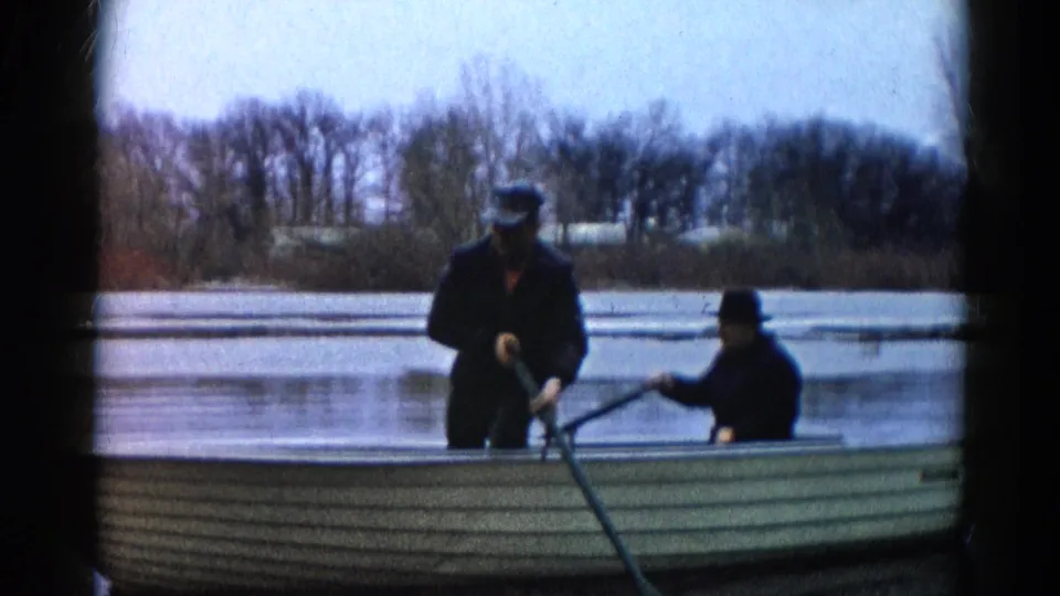 Home-movie frame from Wauconda, Illinois in 1961 showing two men in a rowboat against a quiet lake and winterlike shoreline.