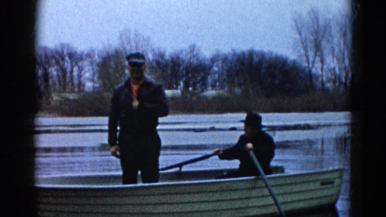 1961 Wauconda, Illinois home-movie frame showing two adult men in a canoe, with one standing near the bow and another seated with a paddle.