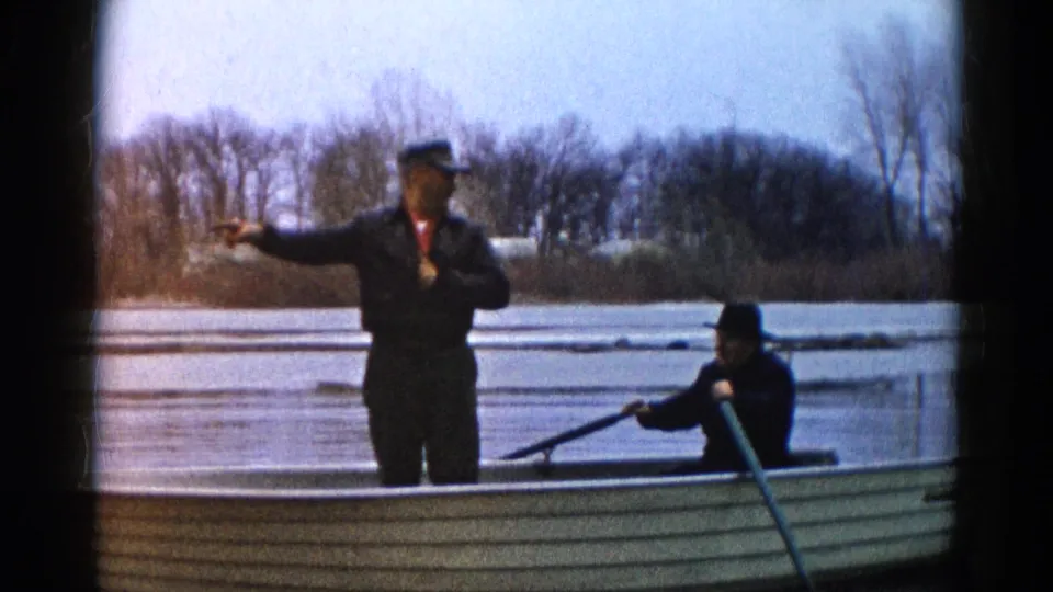 In this 1961 home-movie frame, one man points outward from a canoe while another remains seated with a paddle on the lake.