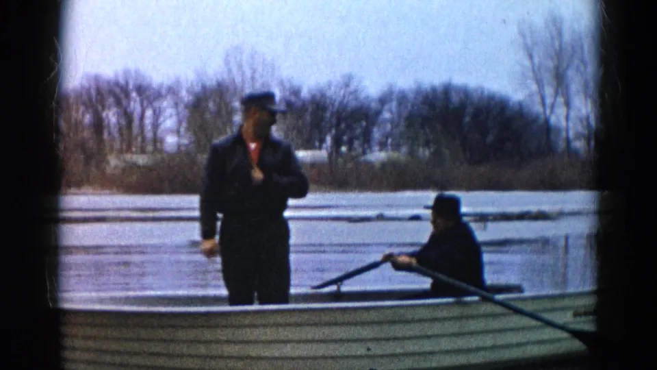 A standing man and seated paddler are framed in a small canoe on a calm Wauconda lake, with bare shoreline trees in the background.