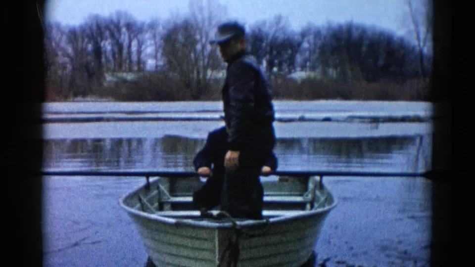 An adult in a cap stands near the stern of a small rowboat on icy water in the supplied 1961 Wauconda setting.