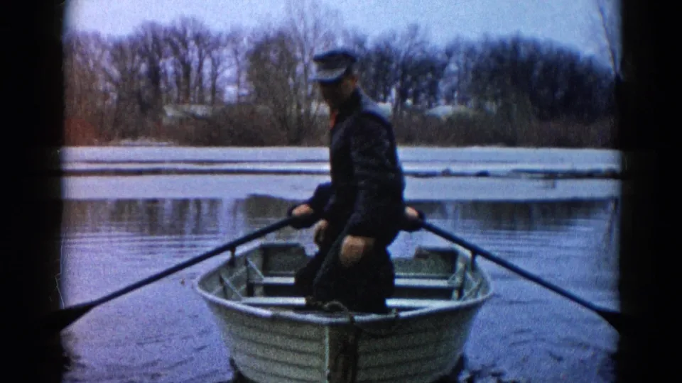 A 1961 Wauconda home-movie frame shows an adult holding an oar in a small aluminum boat on a cold river.