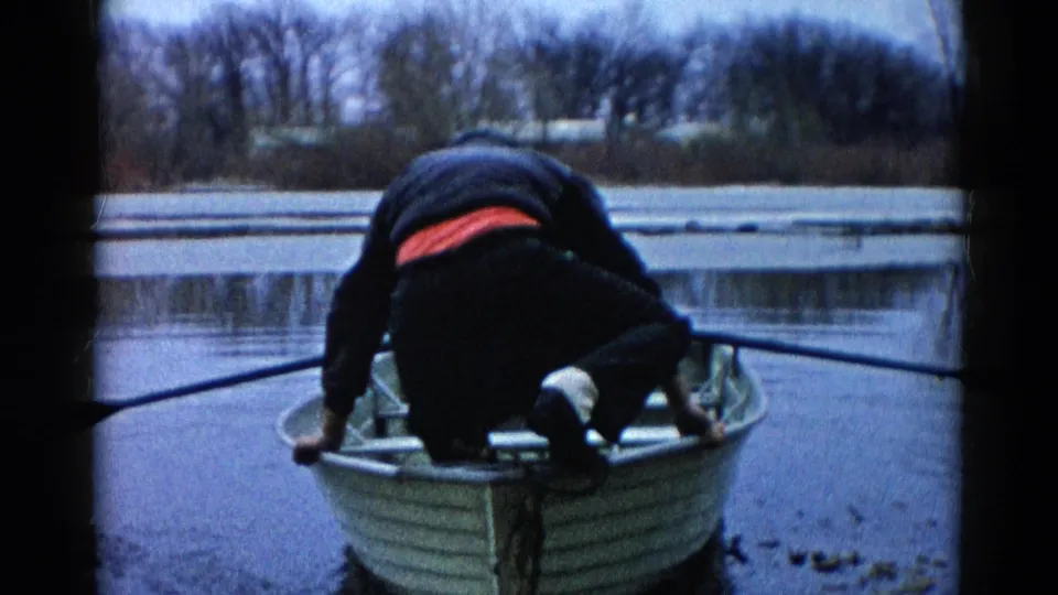 A man crouches low in the aluminum rowboat, with dark winter clothing and a red waistband visible against the muted lake scene.