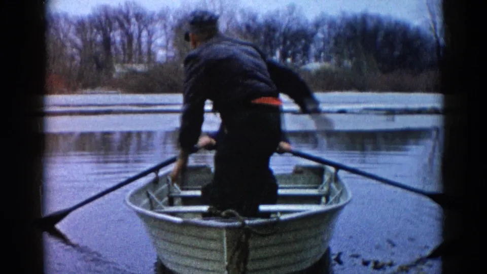 The man regains a more upright position in the small rowboat on the Wauconda lake, with winter trees lining the background.