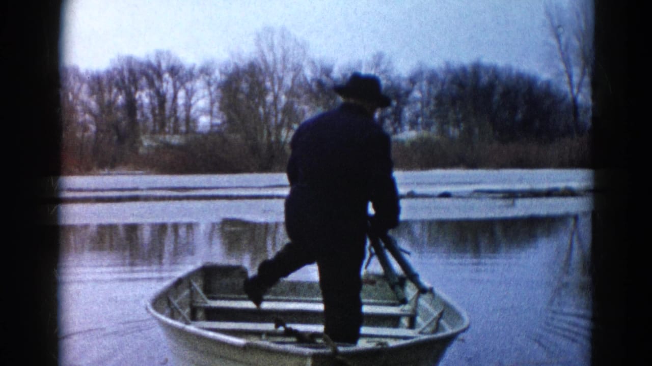 In Wauconda, Illinois, 1961, a man in dark outerwear stands in a small aluminum rowboat on still water near a bare shoreline.