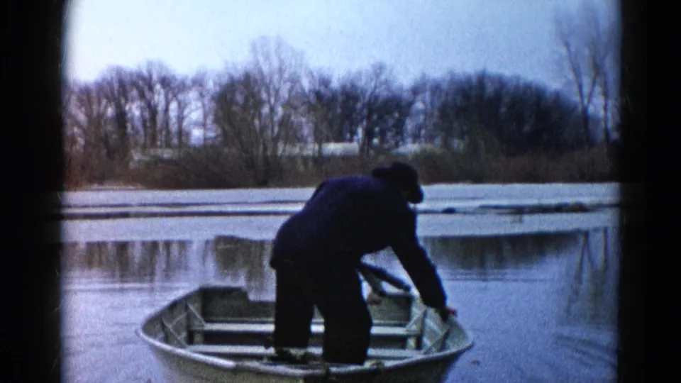 A man bends forward in a small rowboat on a quiet Wauconda lake, with bare trees and pale winter light in the background.