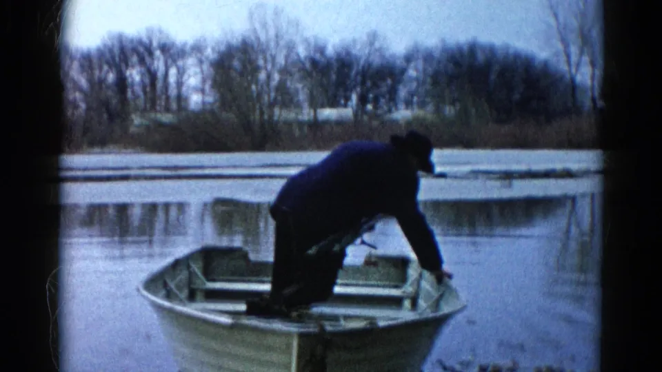 The man crouches low in the aluminum rowboat, reaching toward the gunwale on a calm 1961 Wauconda lake scene.