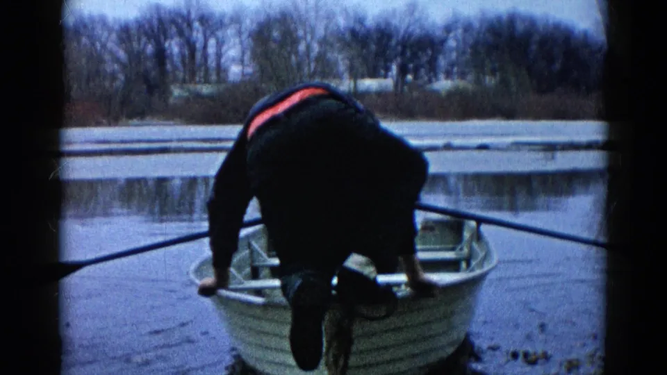 A man steps at the edge of a small rowboat, with long oars visible across the foreground and a bare lakeshore behind him.