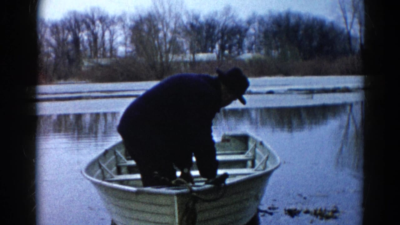 In Wauconda, Illinois, a man in winter clothing leans over gear in an aluminum rowboat on a partly frozen lake in 1961 home-movie footage.