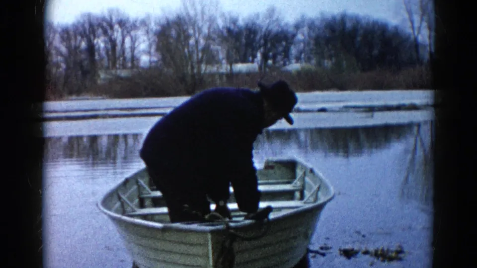 In Wauconda, Illinois, a man in winter clothing leans over gear in an aluminum rowboat on a partly frozen lake in 1961 home-movie footage.