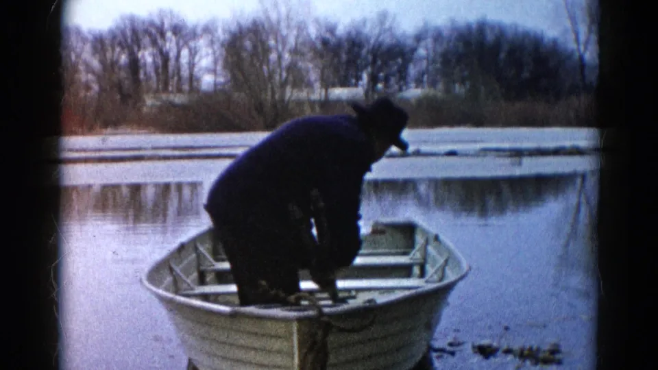 A man in a dark coat and hat leans inside a small aluminum rowboat on a cold Wauconda lake, with ice and winter trees behind him.