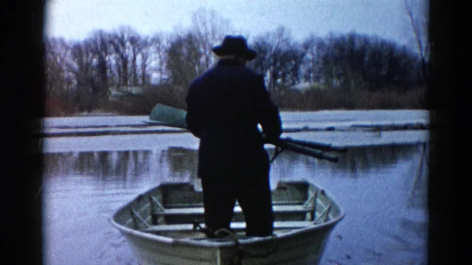 The man stands facing the lake from the rowboat, holding long oars or equipment against a winter shoreline in 1961 Illinois footage.