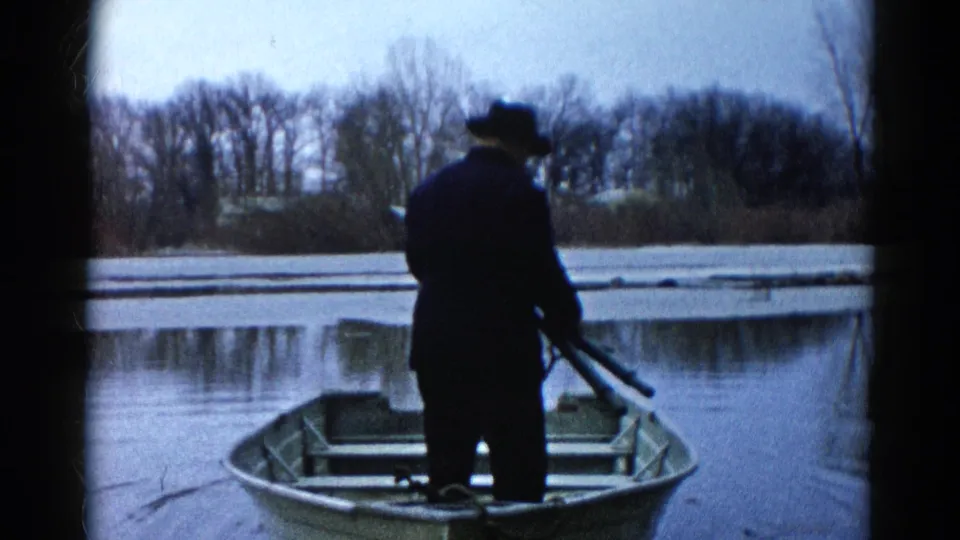 A man in a hat stands in an aluminum rowboat on a calm, partly frozen Wauconda lake, framed by bare winter trees and 8mm film edges.