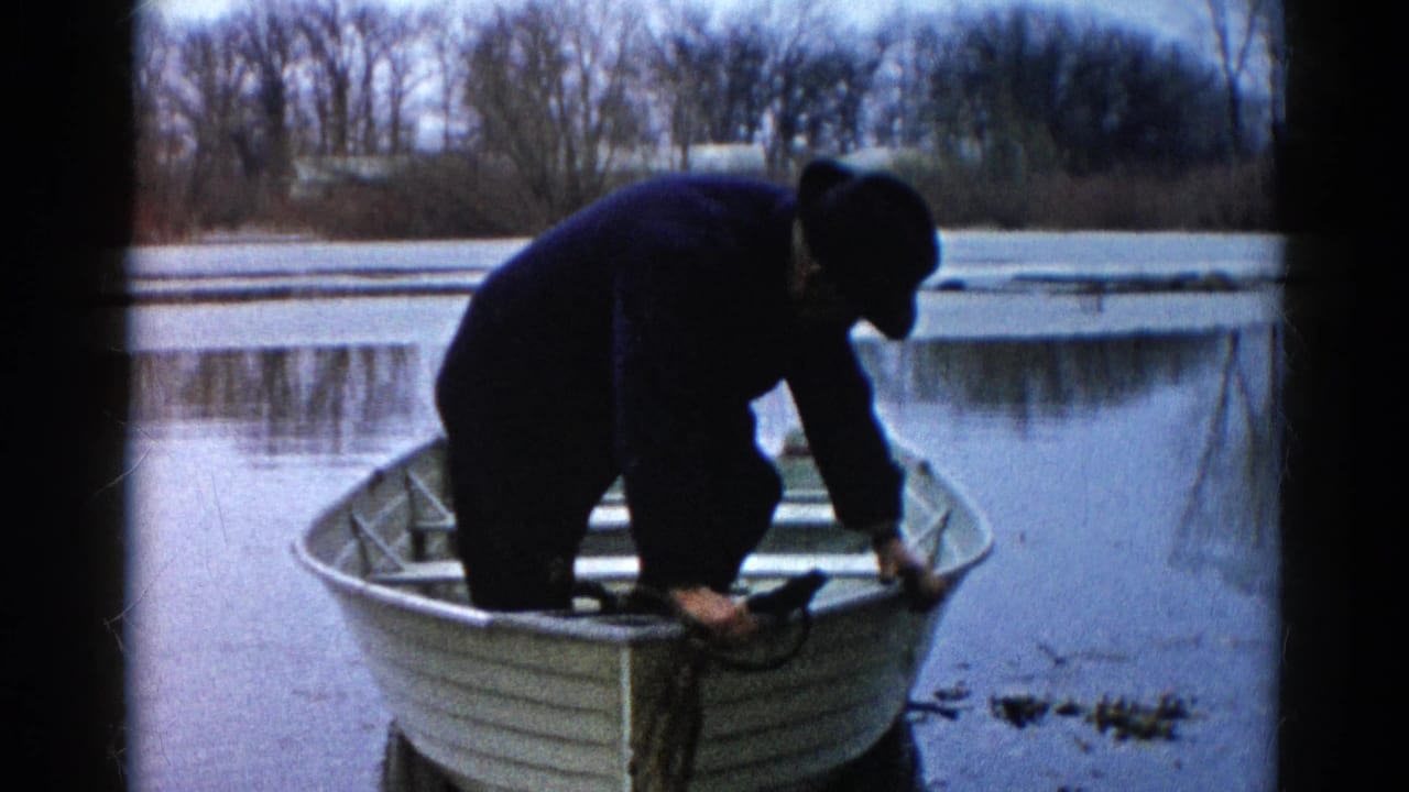 Adult man in Wauconda, Illinois, bends over fishing gear in a small aluminum rowboat on calm water in 1961 home-movie footage.
