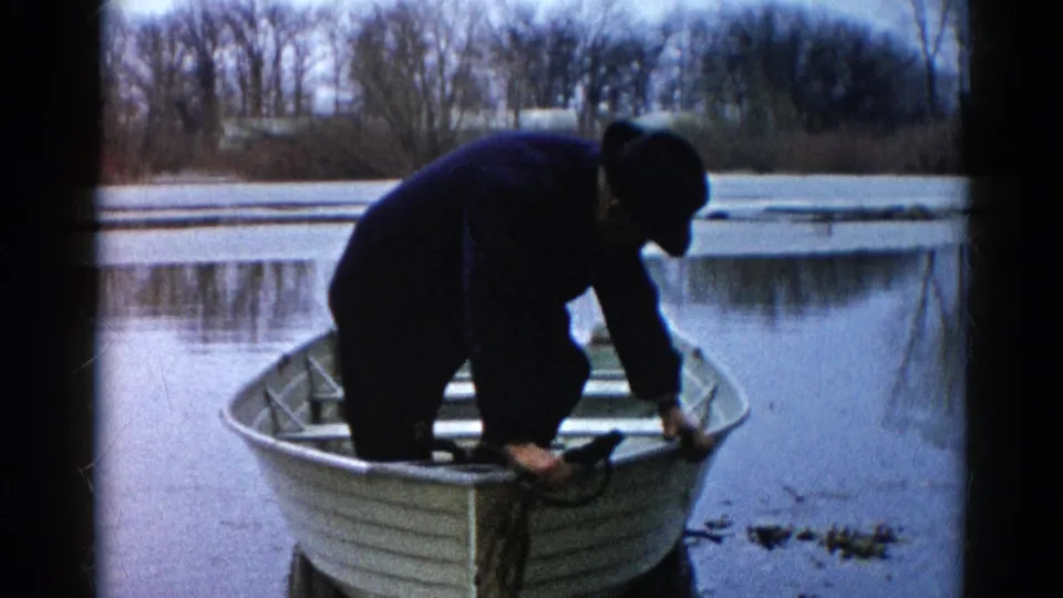 Adult man in Wauconda, Illinois, bends over fishing gear in a small aluminum rowboat on calm water in 1961 home-movie footage.