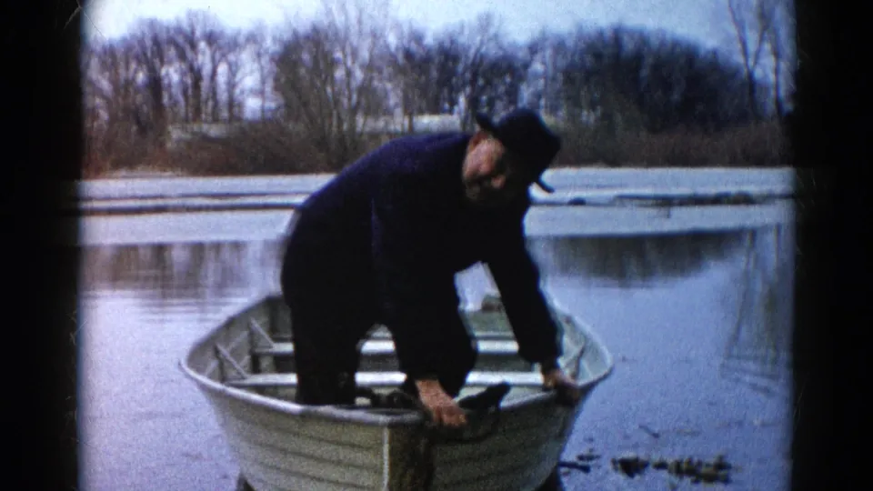 Adult fisherman crouches in a small rowboat on calm Wauconda water, with a bare tree line behind him in 1961 footage.
