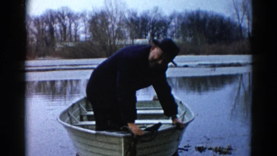 In the 1961 Wauconda home movie, the fisherman bends near the rowboat bow while the still water reflects the wintery shoreline.