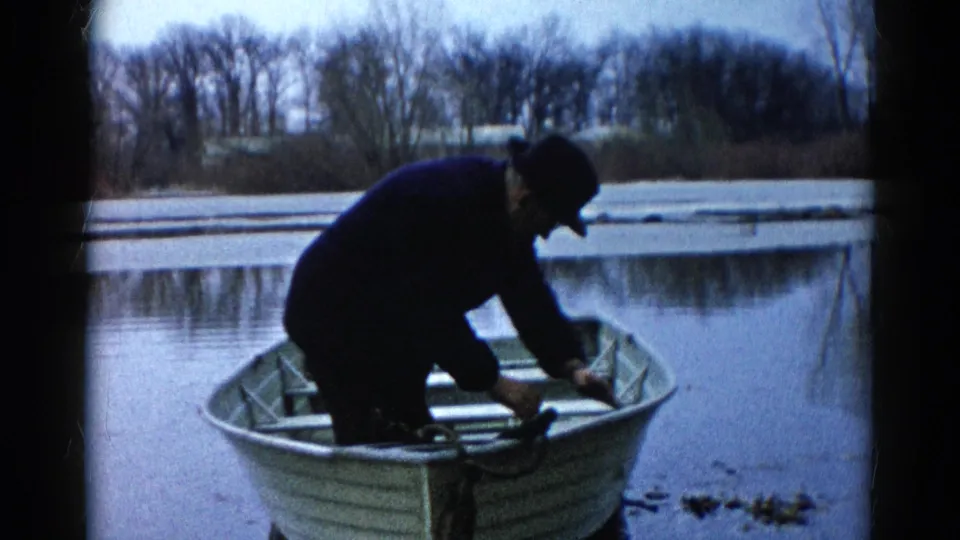 Adult man in dark clothing reaches down inside an aluminum rowboat on Wauconda water, shown in grainy 1961 8mm footage.
