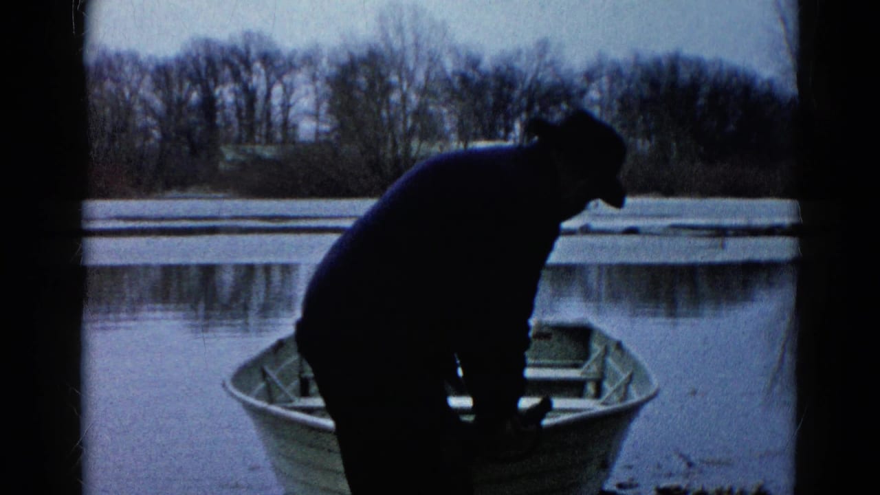 In the 1961 Wauconda home-movie scene, a man in a dark coat leans toward a light-colored rowboat beside a calm river.