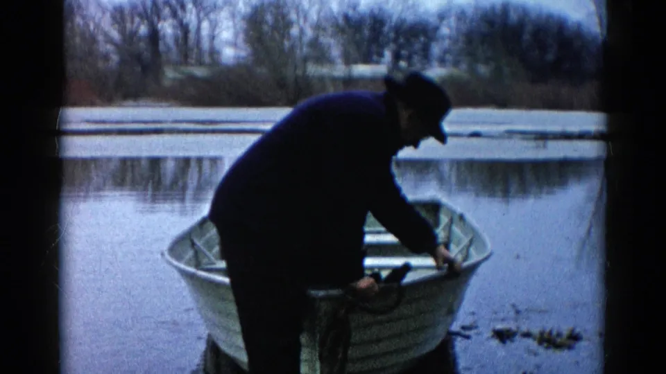 A man in dark clothing steadies himself at the bow area of a rowboat on a calm river in Wauconda, Illinois.