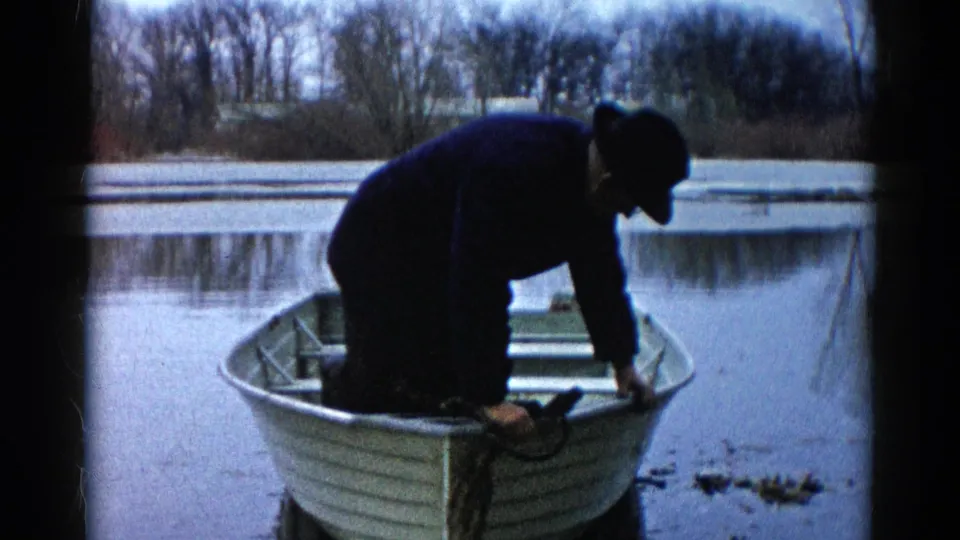 The man appears partly inside the rowboat, crouched low as he adjusts his footing near the calm riverbank.