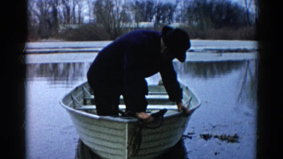 A man in a dark coat bends forward inside the rowboat, framed against still water and a leafless riverside background.